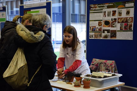 Eine Teilnehmerin des Schülerforschungszentrums Kieler Forschungswerkstatt präsentiert ihr Projekt über nachhaltiges Bio-Baumaterial auf einem Wettbewerb, während sie mit interessierten Besucherinnen am Stand spricht und ihr Material demonstriert.